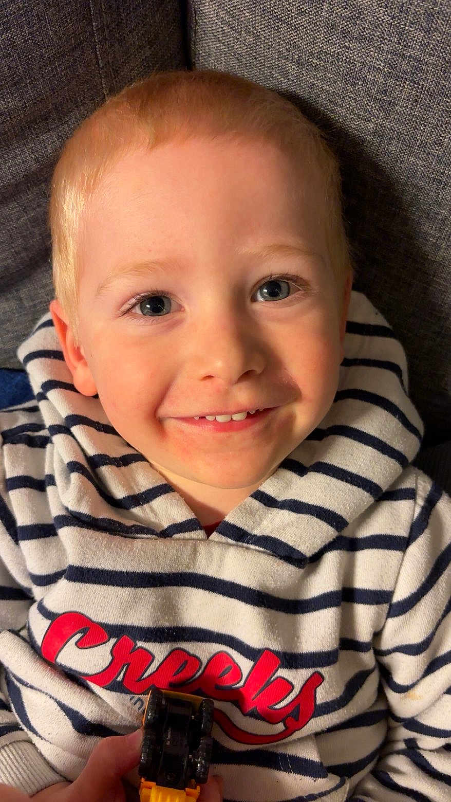 Charly a rejoint le concours — aidez-le/la à gagner de superbes lots ! child, smiling, blue_eyes, striped_hoodie, toy_vehicle, yellow_toy, close_up, portrait, happy, indoors, couch, fabric, casual_clothing, playful, young_child, human, face, person, cute, kid