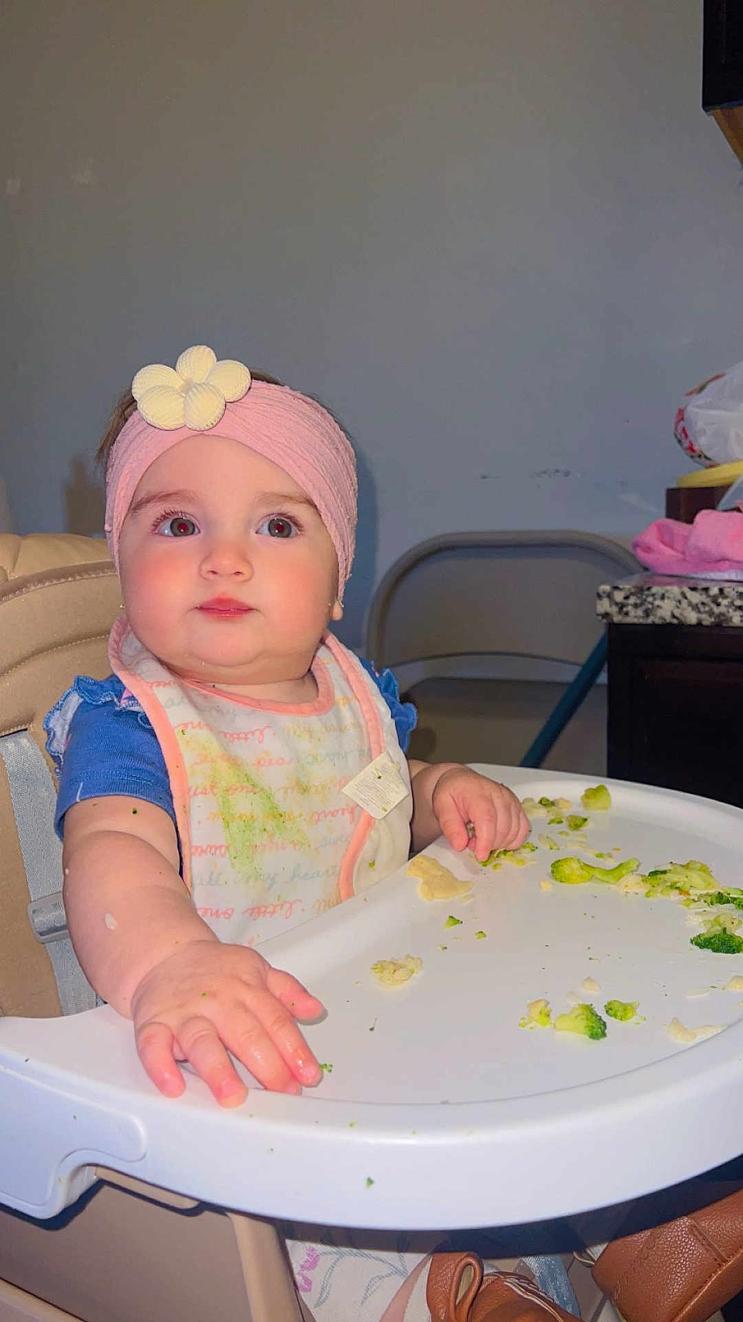 Khloe joined the competition — help win amazing prizes! baby, child, high_chair, pink_headband, bib, food, broccoli, indoor, curious, cute, infant, table, feeding, seat, clothing, face, hand, expression, kitchen, furniture
