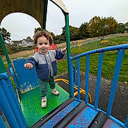 Theo joined the competition — help win amazing prizes! casual_clothing, child, curly_hair, fence, grass, greenery, metal, outdoor, overcast_sky, park, person, play_equipment, playground, shoes, slide, smiling, sweater, toddler, trees, young_child