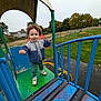 casual_clothing, child, curly_hair, fence, grass, greenery, metal, outdoor, overcast_sky, park, person, play_equipment, playground, shoes, slide, smiling, sweater, toddler, trees, young_child