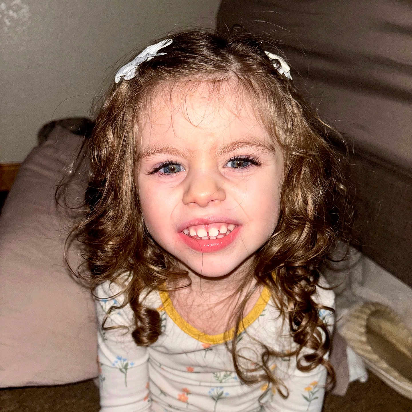 Lainey is registered to the contest to win money with this photo: carpet, child, closeup, cozy, curly_hair, cushion, face, girl, hair_clip, happy, indoor, long_sleeve, pajamas, patterned_clothing, person, portrait, seated, slippers, smiling, young
