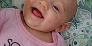 Sunshine is registered to the contest to win money with this photo: baby, smiling, face, person, infant, clothing, pink_shirt, text_on_clothing, quilt, pattern, happy, portrait, closeup, child, cute, skin, head, lying_down, indoor, expression