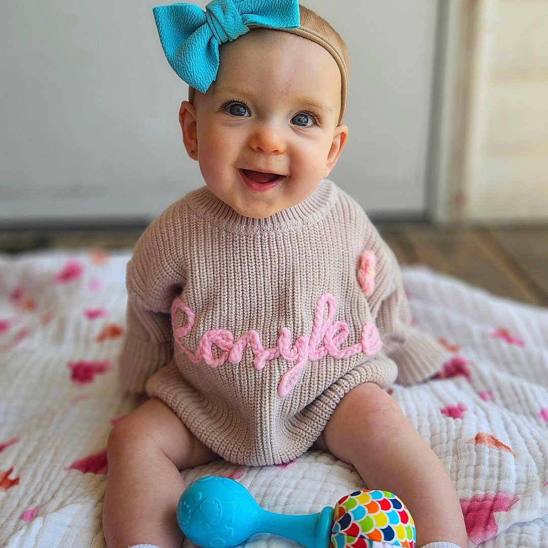 Rosylee is registered to the contest to win money with this photo: baby, blanket, blue_bow, candid, child, colorful_toy, cozy, cute, floor, happy, headband, indoor, infant, knitted_sweater, playful, portrait, sitting, smiling, sock, toy