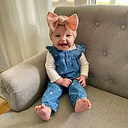 Lucia joined the competition — help win amazing prizes! baby, child, smiling, headband, bow, denim, overalls, sitting, gray_chair, indoor, happy, cute, infant, person, portrait, feet, window, curtains, clothing, home
