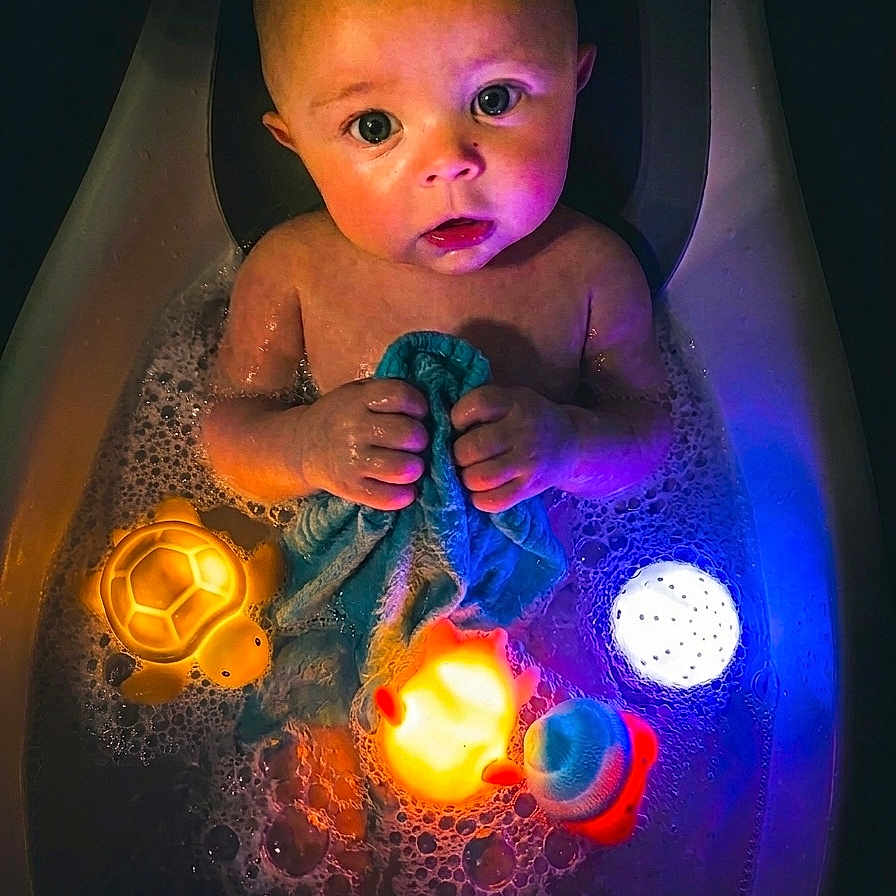 Rowdy is registered to the contest to win money with this photo: baby, bathtub, bathwater, bubbles, child, toy, bath_toy, glowing, blue, orange, yellow, water, skin, cloth, cute, indoors, head, face, sitting, expression