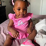 Mahogany is registered to the contest to win money with this photo: baby, child, pink_dress, pink_shoes, bows, hair_accessories, sitting, bed, blanket, indoor, cute, portrait, infant, happy, clothing, footwear, person, young_child, home, soft_light