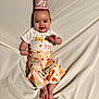 baby, infant, crown, half_birthday, smiling, floral_pants, white_blanket, cute, celebration, lying_down, child, happy, portrait, clothing, footwear, headwear, indoors, person, infant_fashion, milestone