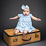 baby, child, blue_dress, bow, sitting, vintage_suitcase, studio, portrait, cute, infant, smiling, barefoot, expression, isolated, gray_background, person, young_child, studio_lighting, fashion, accessory