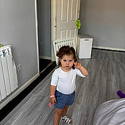 Amela is registered to the contest to win money with this photo: toddler, child, indoor, standing, curly_hair, white_top, blue_shorts, sandals, floor, door, chair, keychain, hand_gesture, serious_expression, home, radiator, plastic_container, wall, person, portrait