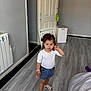 toddler, child, indoor, standing, curly_hair, white_top, blue_shorts, sandals, floor, door, chair, keychain, hand_gesture, serious_expression, home, radiator, plastic_container, wall, person, portrait