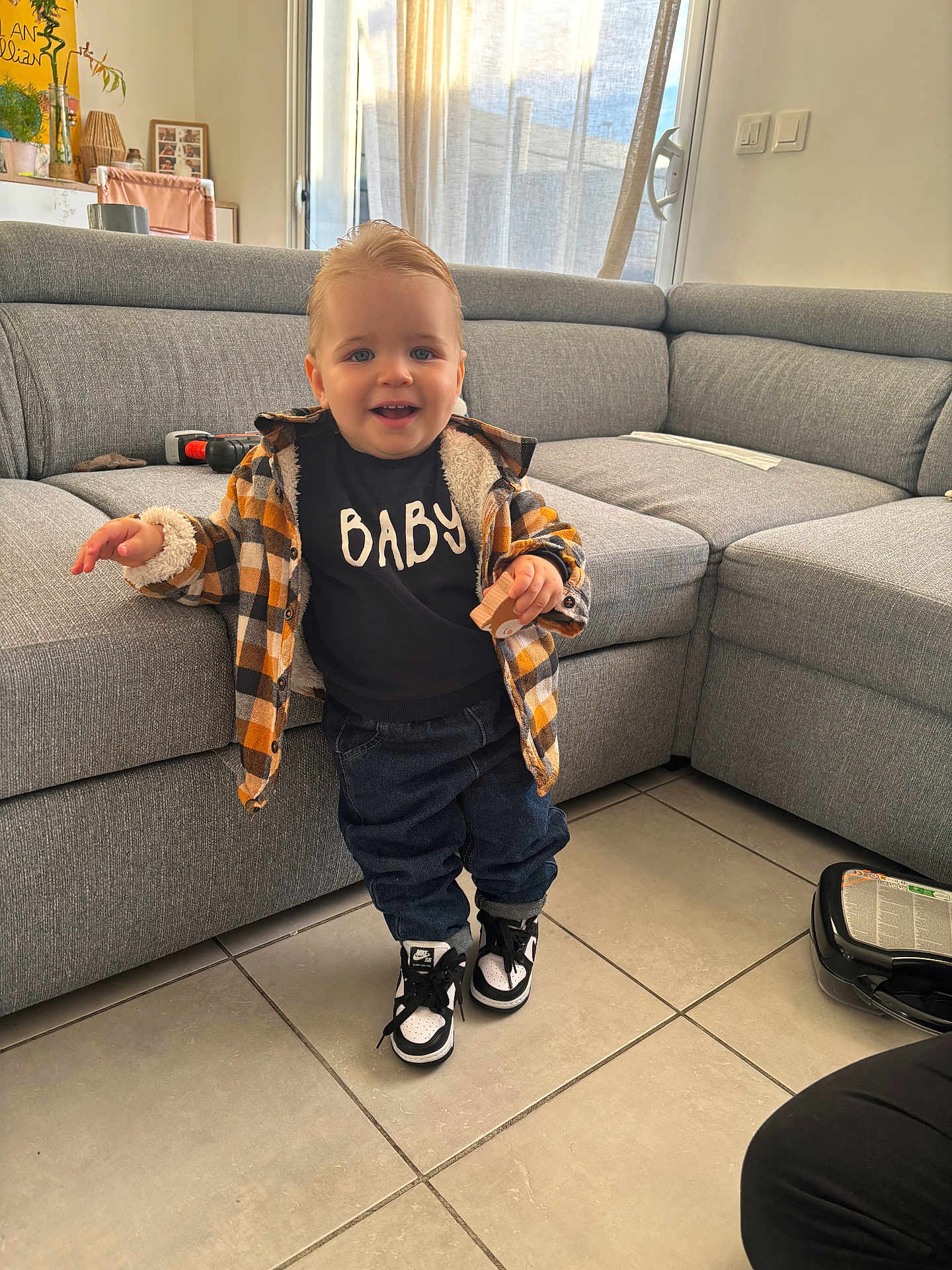 Giullian participe au concours pour gagner de l'argent avec cette photo : toddler, child, indoor, living_room, plaid_jacket, sneakers, toy, smiling, curtains, couch, floor_tiles, window, person, clothing, happy, hair, furniture, home, casual, portrait