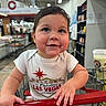 Leonidas joined the competition — help win amazing prizes! baby, child, shopping_cart, store, aisle, shelf, clothing, onesie, hand, face, smile, cheeks, indoor, person, retail, grocery, curly_hair, red_handle, drink, shopping