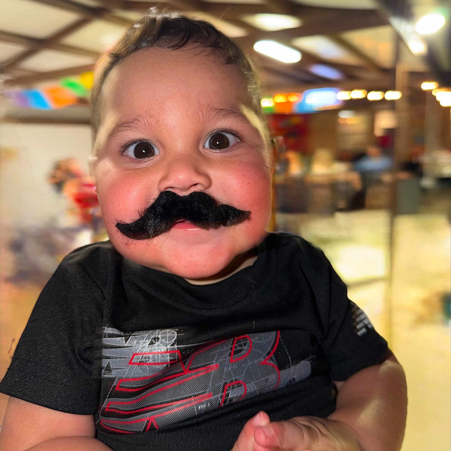 Leonidas is registered to the contest to win money with this photo: toddler, child, mustache, black_shirt, indoor, portrait, cute, funny, expression, hands, smile, face, person, young_child, black_hair, clothing, blurred_background, warm_lighting, closeup, playful