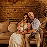 family, baby, newborn, mother, father, couch, brick_wall, indoor, portrait, smile, dress, beige, wooden_floor, pampas_grass, decor, intimate, warm_lighting, seated, happy, people