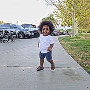 Kaden is registered to the contest to win money with this photo: toddler, child, smiling, happy, outdoor, sidewalk, parked_cars, trees, grass, portrait, standing, walking, curly_hair, afro_hair, white_shirt, blue_shorts, sandals, daylight, roadside, vehicles
