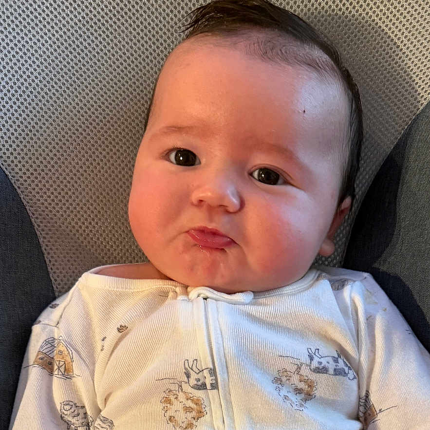 Daniel is registered to the contest to win money with this photo: baby, infant, face, pout, onesie, clothing, cute, child, portrait, sitting, indoors, expression, head, skin, hair, young, newborn, person, texture, background