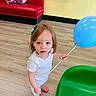toddler, child, balloon, blue_balloon, hair_clip, pink_shoes, white_clothing, indoor, play_area, wooden_floor, green_chair, red_bench, curious_expression, person, young_child, flooring, toy, casual_clothing, looking_up, standing