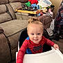 Ronnie is registered to the contest to win money with this photo: toddler, child, high_chair, spiderman_outfit, smiling, indoor, living_room, couch, toys, clothes, floor, wooden_floor, face, person, furniture, home, cute, young_child, happy, seated