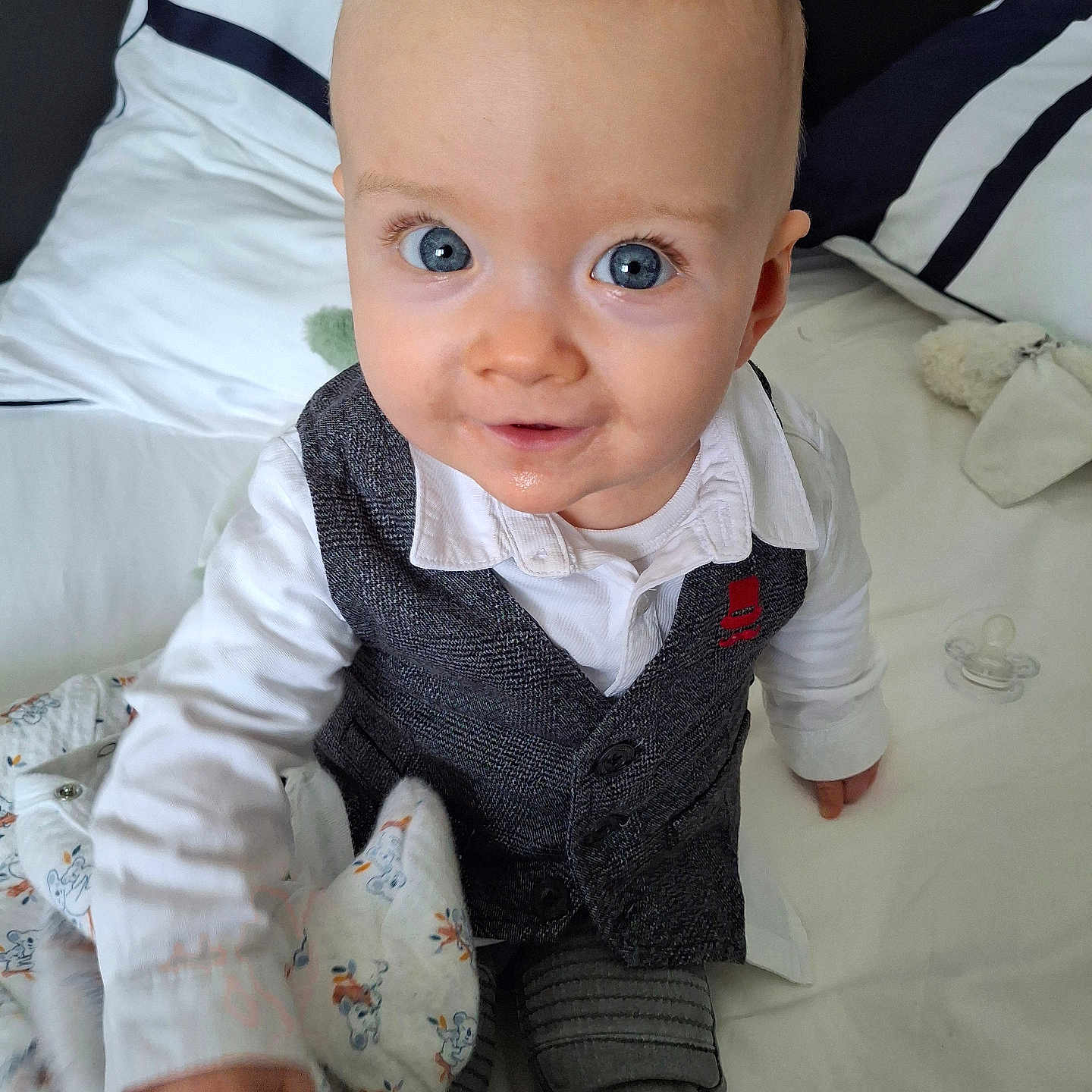 Eden participe au concours pour gagner de l'argent avec cette photo : baby, bed, blanket, blue_eyes, child, clothing, cozy, curious, cute, face, indoors, infant, pacifier, person, pillow, portrait, sitting, smile, vest, white_shirt