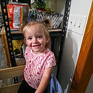 Riley is registered to the contest to win money with this photo: child, toddler, smiling, pigtails, indoor, kitchen, snacks, pantry, shelf, light_switch, wooden_doorframe, clothing, pink_shirt, black_pants, person, happy, standing, handrail, face, hair