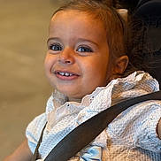 Esmée participe au concours pour gagner de l'argent avec cette photo : toddler, child, smiling, face, blue_eyes, white_dress, bow, car_seat, seatbelt, closeup, portrait, happy, cute, young_child, baby, indoor, clothing, person, skin, hair