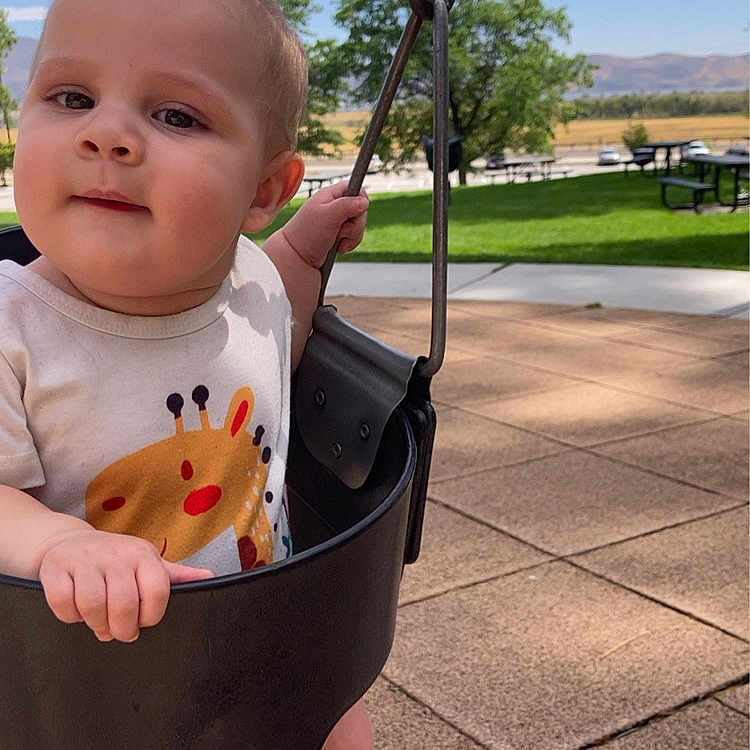 Kyle is registered to the contest to win money with this photo: baby, child, infant, swing, playground, outdoor, park, smiling, portrait, face, person, sitting, standing, sunlight, blue_sky, mountains, trees, grass, pavement, playground_equipment