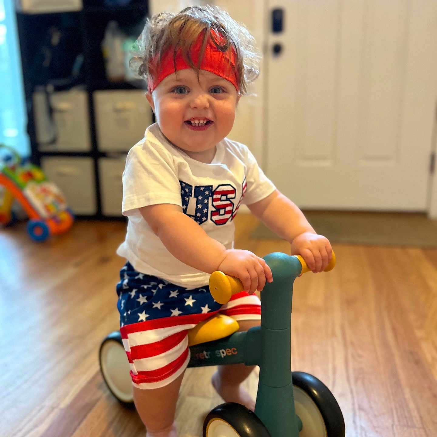 Levi is registered to the contest to win money with this photo: baby, clothing, face, happy, hardwood, head, indoors, machine, pants, person, photography, portrait, shorts, smile, t_shirt, transportation, tricycle, vehicle, wheel, wood