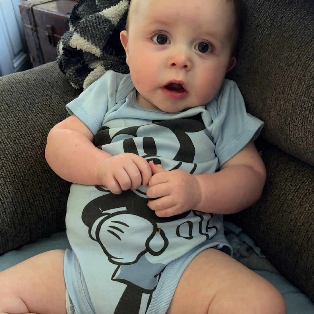 Darryl is registered to the contest to win money with this photo: baby, child, infant, sitting, armchair, couch, blanket, cartoon_clothing, blue_outfit, portrait, indoors, home, wide_eyed, open_mouth, smiling, hands, legs, bare_feet, vintage_trunk, cozy