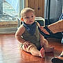 baby, child, indoor, floor, wooden_floor, romper, bib, footwear, sandal, toenails, window, wooden_wall, person, sitting, skin, hand, face, expression, light, home