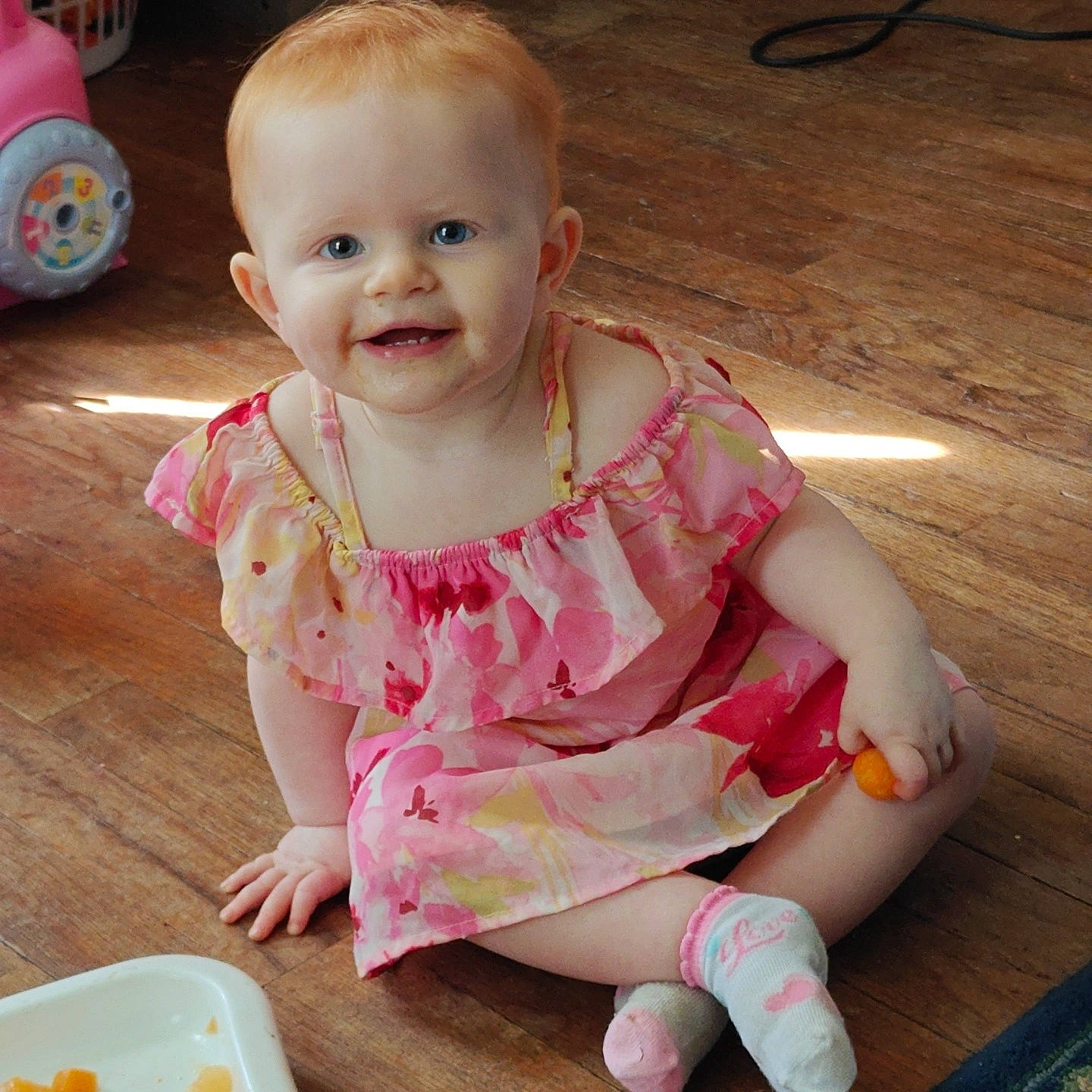 Eliza is registered to the contest to win money with this photo: baby, baby_toddler_clothing, cheek, child, dress, flooring, fun, happy, hardwood, human_leg, iris, joy, pattern, person, pink, sitting, skin, sleeve, smile, tableware