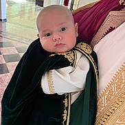 Brayan-Adam a rejoint le concours — aidez-le/la à gagner de superbes lots ! baby, child, person, traditional_clothing, embroidery, portrait, indoor, holding, face, curious, chubby_cheeks, head, black_clothing, gold_detail, people, background, floor, light, wall, door