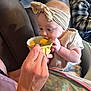 baby, infant, feeding, spoon, bowl, headband, bib, highchair, adult_hand, blanket, couch, indoor, portrait, puree_food, sitting, closeup, clothing, person, plaid_jacket, tattooed_hand