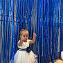 Léna a rejoint le concours — aidez-le/la à gagner de superbes lots ! balloon, blue_accent, blue_fringe, celebration, child, cute, dress, event, floor, fringe_backdrop, fun, indoor, party, person, playful, pose, smiling, toddler, white_dress, young_child