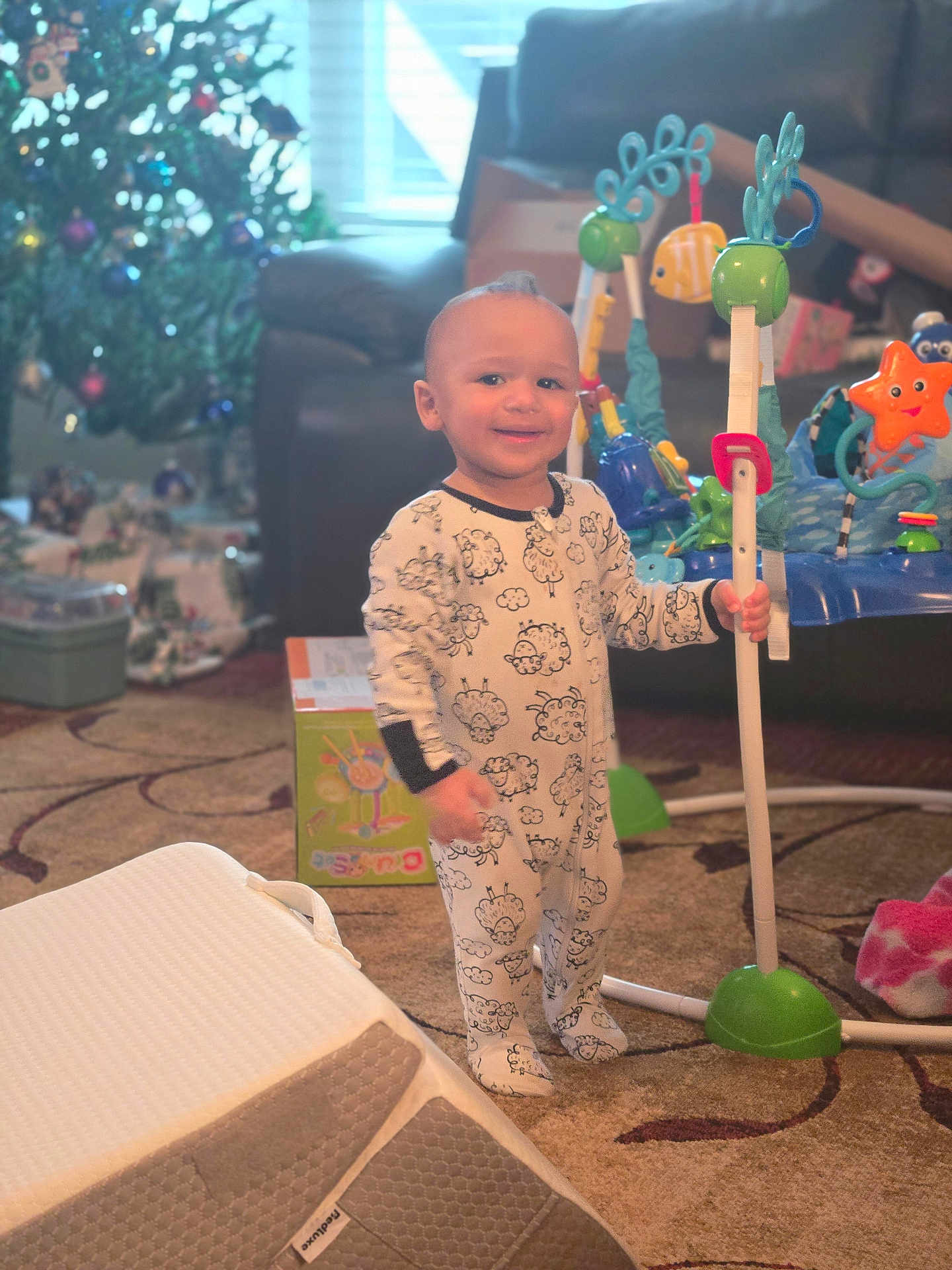 Cameron joined the competition — help win amazing prizes! toddler, child, pajamas, sheep_pattern, baby_toys, activity_center, christmas_tree, living_room, carpet, sofa, holiday_decor, smiling, indoors, toy_box, baby, person, playtime, festive, home, cute