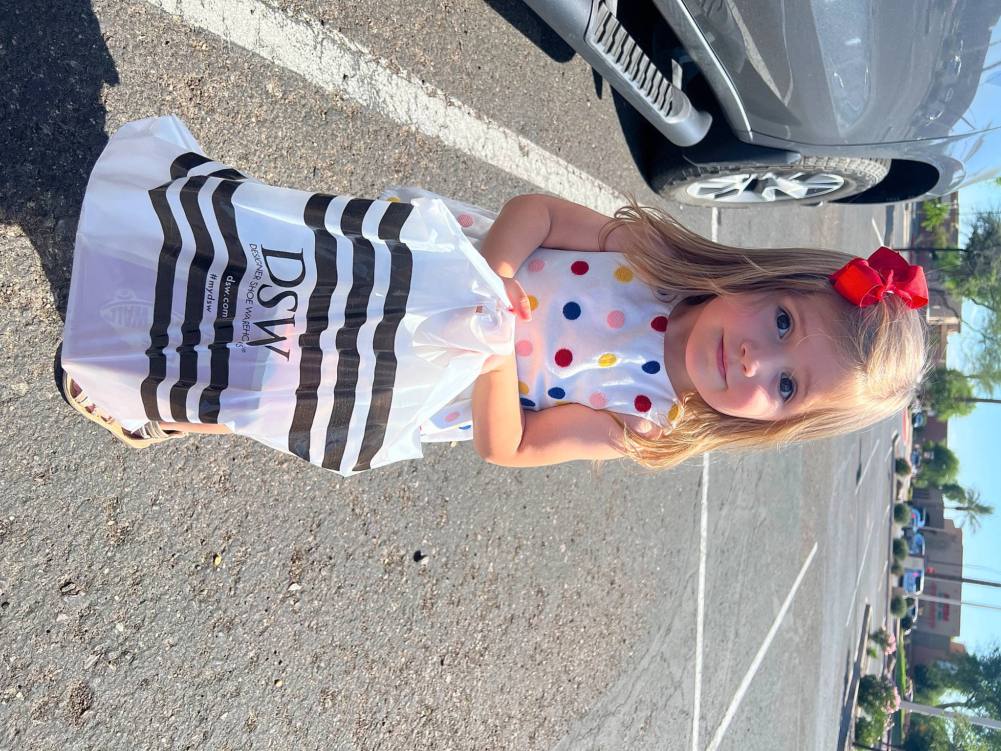 Elle is registered to the contest to win money with this photo: asphalt, auto_part, automotive_exterior, automotive_lighting, automotive_tire, bumper, car, fun, hat, human_leg, joy, motor_vehicle, person, sidewalk, sky, tire, toddler, tree, trunk, vehicle