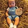 baby, infant, child, overalls, socks, hay, nest, sign, four_months, portrait, cute, sitting, holding, expression, warm_lighting, outdoor, nature, closeup, soft_skin, small_hand