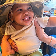 Donico-Juan is registered to the contest to win money with this photo: baby, child, smile, hat, bucket_hat, hands, person, indoor, clothing, green_pants, white_shirt, happy, cute, portrait, infant, face, skin, fingers, bracelet, tattoo