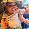 baby, child, smile, hat, bucket_hat, hands, person, indoor, clothing, green_pants, white_shirt, happy, cute, portrait, infant, face, skin, fingers, bracelet, tattoo