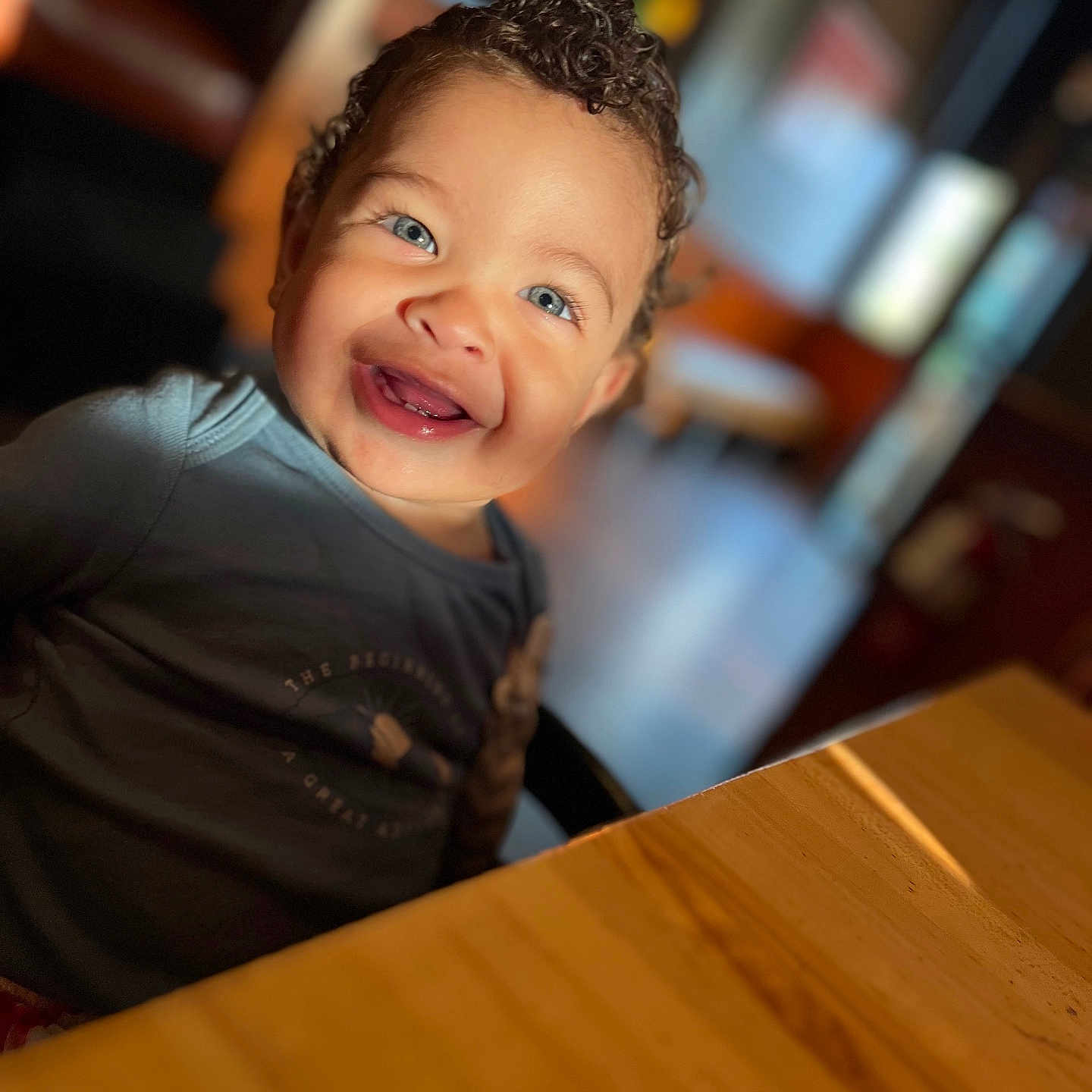 Jac joined the competition — help win amazing prizes! background_blur, blue_eyes, casual_clothing, cheerful, child, curly_hair, cute, face, happy, indoor, natural_light, person, portrait, seated, smiling, table, toddler, warm_lighting, wood, young