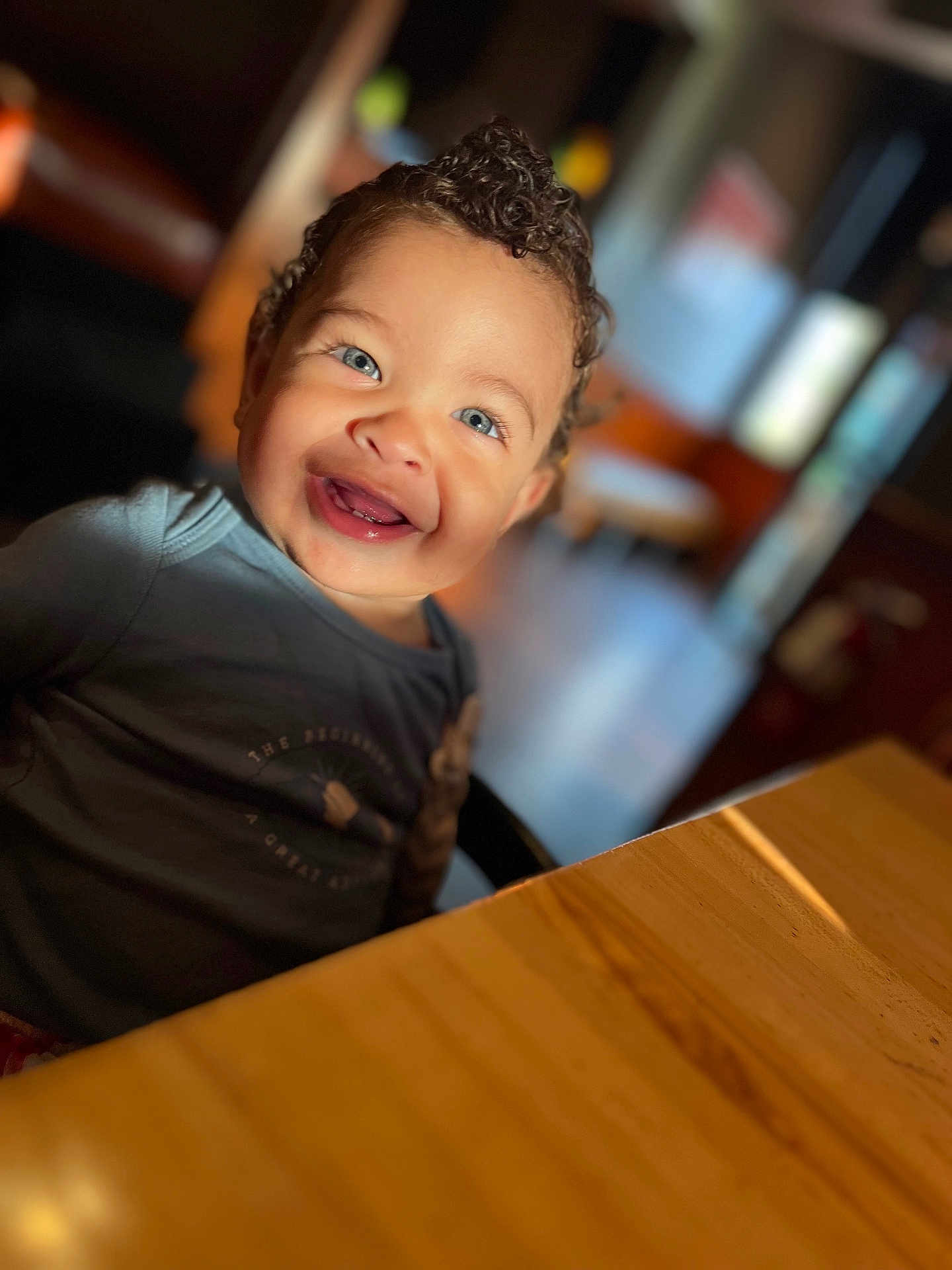 Jac joined the competition — help win amazing prizes! child, toddler, smiling, blue_eyes, curly_hair, happy, portrait, indoor, table, wood, casual_clothing, face, person, cute, young, seated, warm_lighting, background_blur, natural_light, cheerful