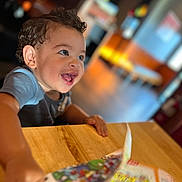 Jac joined the competition — help win amazing prizes! child, toddler, curly_hair, happy, smiling, indoor, table, wooden_table, book, magazine, colorful, reaching, face, person, blurred_background, bright_eyes, excited, casual_clothing, young_child, playful