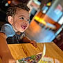 child, toddler, curly_hair, happy, smiling, indoor, table, wooden_table, book, magazine, colorful, reaching, face, person, blurred_background, bright_eyes, excited, casual_clothing, young_child, playful