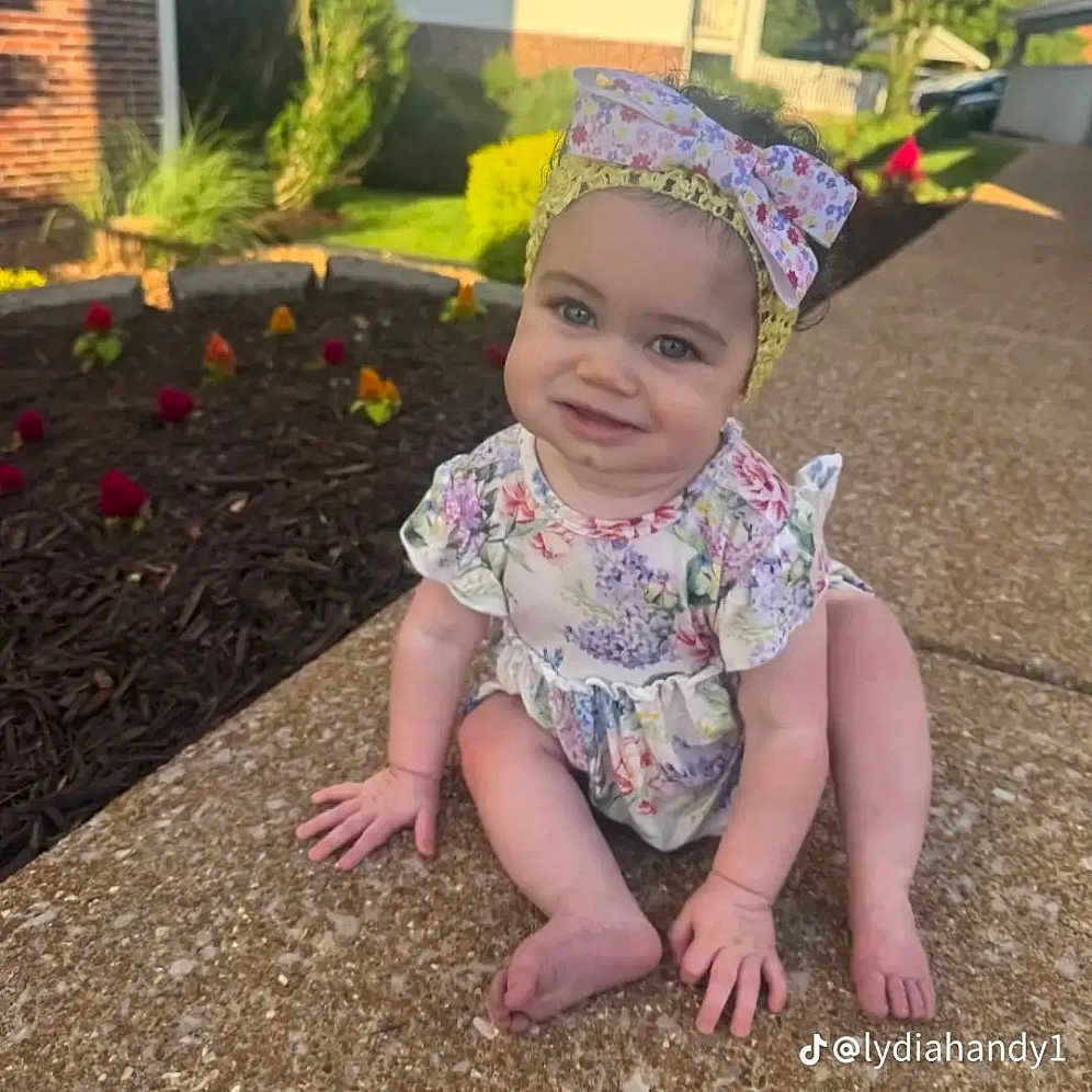 Amelia is registered to the contest to win money with this photo: baby, child, infant, sitting, outdoors, sidewalk, garden, flowers, floral_outfit, headband, bow, smiling, face, portrait, suburban, house, sunlight, pavement, social_media_watermark, happy