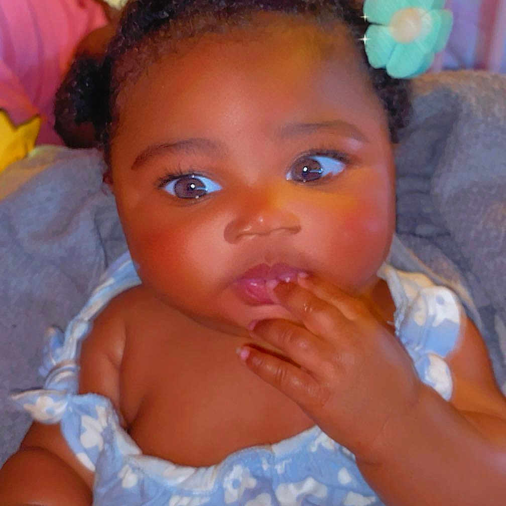 Jalinah Miller is registered to the contest to win money with this photo: adorable, baby, blue_flower, child, closeup, curly_hair, cute, dress, expression, face, fingers_in_mouth, flower_pattern, hair_accessories, hand, indoor, infant, pink_bow, portrait, seated, skin
