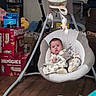 baby, baby_swing, infant, indoor, floor, diapers, boxes, scooter, toy, plush_toy, wooden_floor, person, clothing, footwear, face, expression, seat, blanket, comfort, home