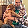affection, beard, brown_dog, casual_clothing, comfort, couch, curtains, dog, fluffy, home, human_animal_interaction, indoor, man, pet, plaid_shirt, portrait, relaxed, seated, warm_lighting, window