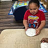 activity, books, child, classroom, cotton_balls, curly_hair, educational_rug, furniture, hand, indoor, learning, person, plate, playroom, shelf, sitting, spiderman_tshirt, table, toddler, wood_floor