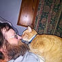 man, beard, glasses, orange_cat, cat_collar, indoor, curtains, wood_panel, bed, blanket, closeup, face, pet, animal, human, cozy, interaction, feline, comfort, resting
