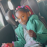 Summer is registered to the contest to win money with this photo: braids, car_interior, car_seat, casual_clothing, child, colorful_beads, girl, green_jacket, happy, holding_object, indoor, person, playful, portrait, seatbelt, sitting, smile, toy, window, young_child
