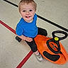 Kasen is registered to the contest to win money with this photo: toddler, child, toy_car, indoor, floor, blue_shirt, black_pants, white_shoes, smiling, happy, play, steering_wheel, plastic_toy, young_child, person, floor_lines, recreation, casual_clothing, cute, active